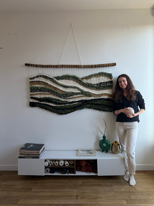 Artist standing next to the handwoven tapestry “Whispers of the Jungle” in green and cream tones