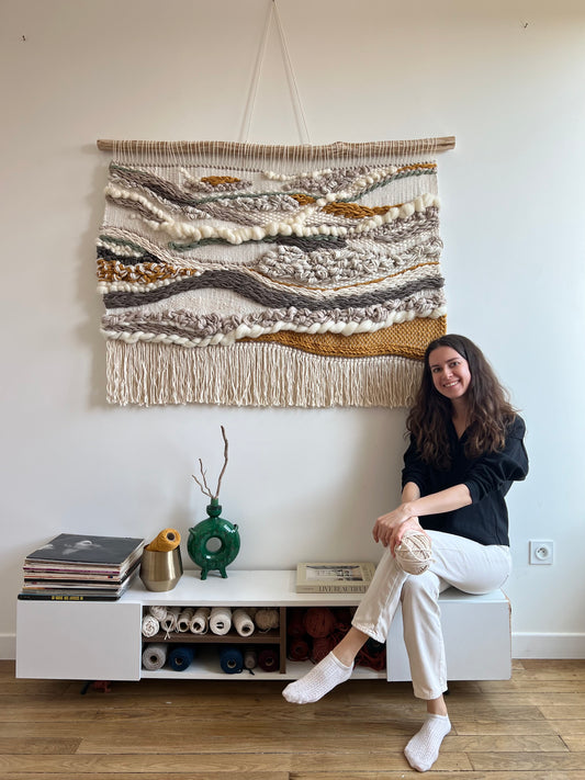 Artist sitting next to large 3D wall tapestry in neutral tones
