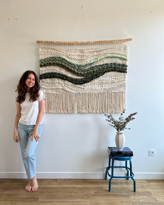 Artist with woven wall hanging with green waves on cream background, unique fiber art tapestry