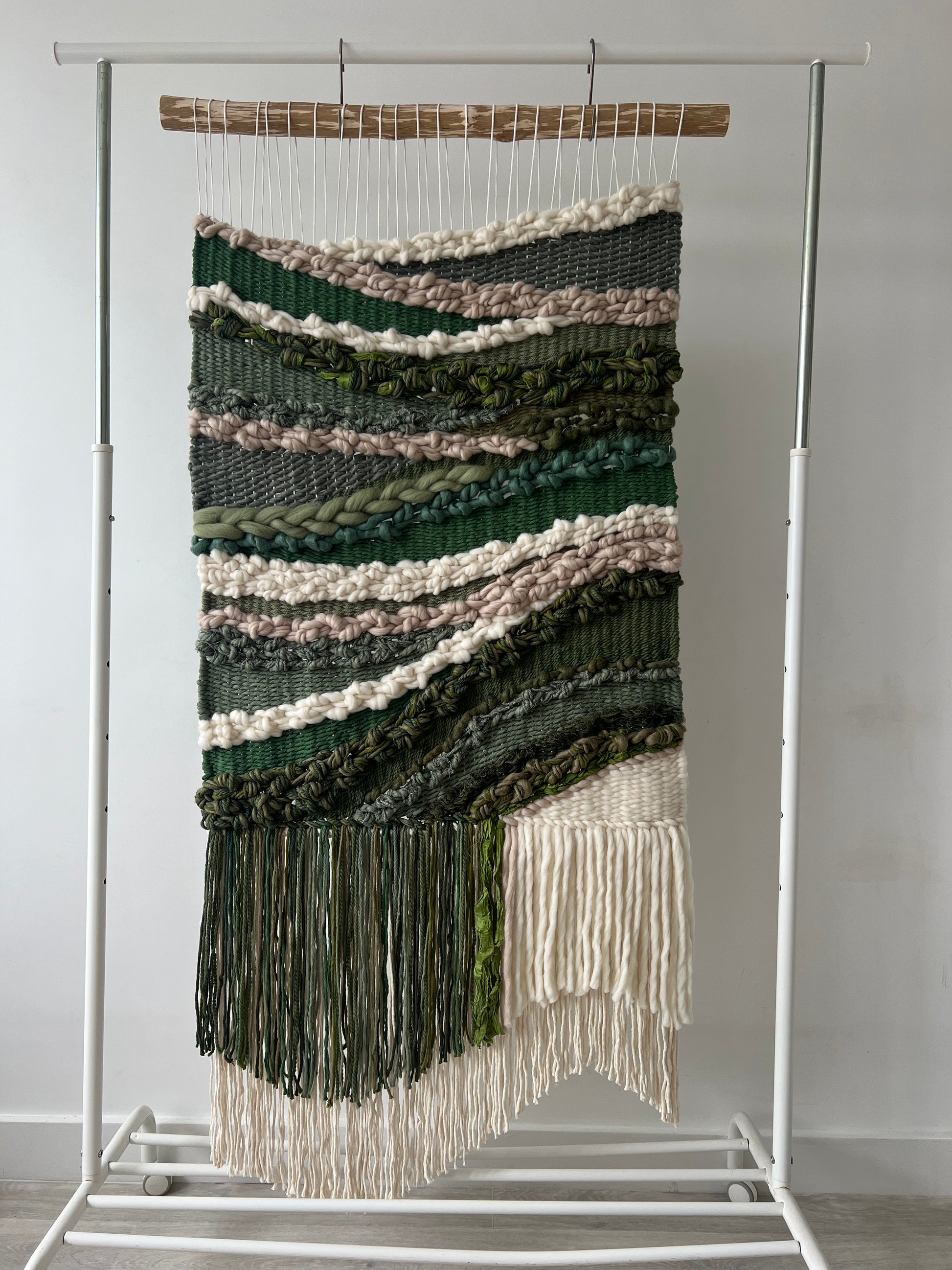 Handwoven wool and cotton wall hanging in earthy green, beige, and cream tones, displayed on a wooden dowel against a white wall.