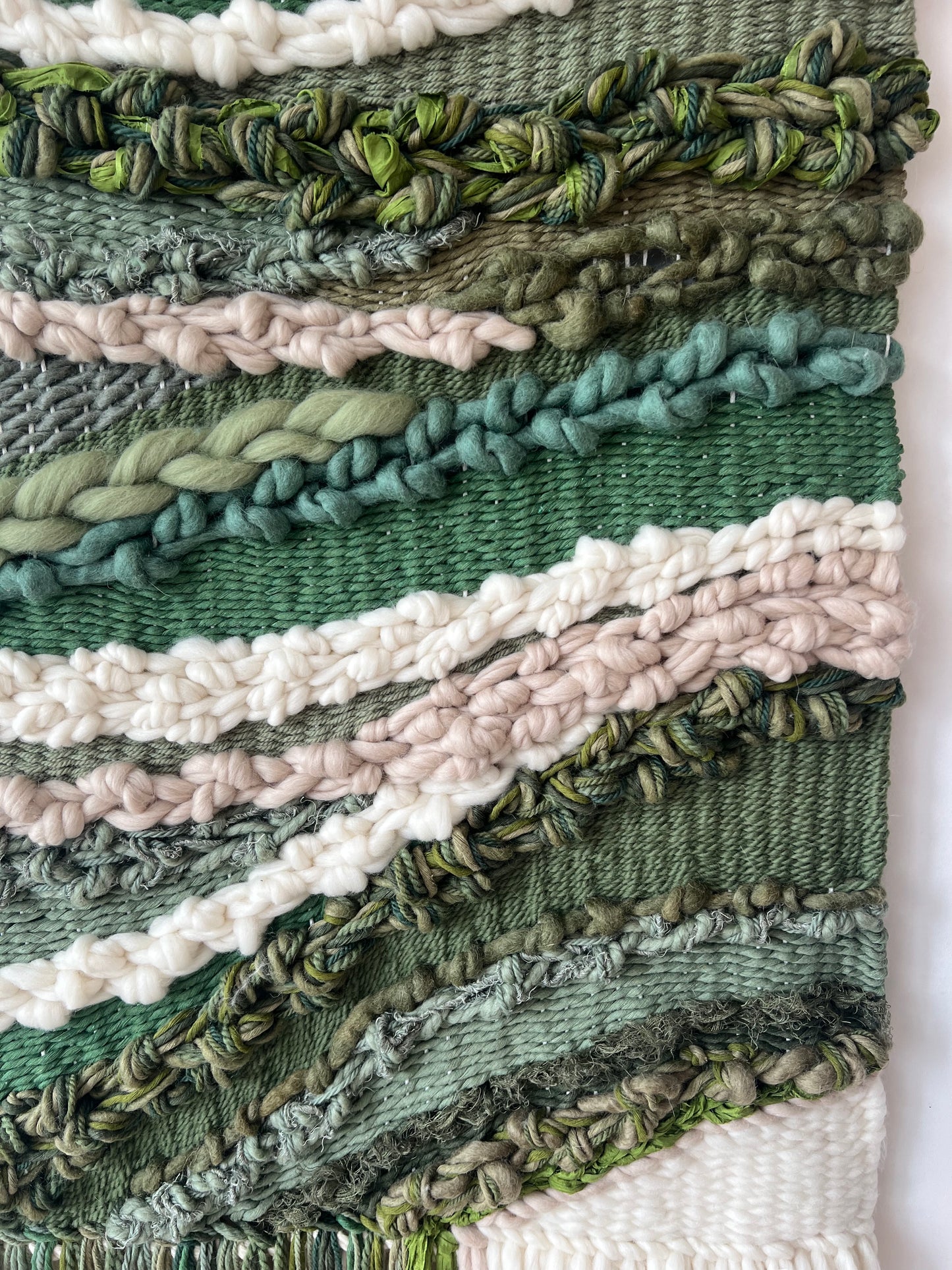 Close-up of soft wool fibers in green, cream and beige tones, showing woven texture of the wall hanging.