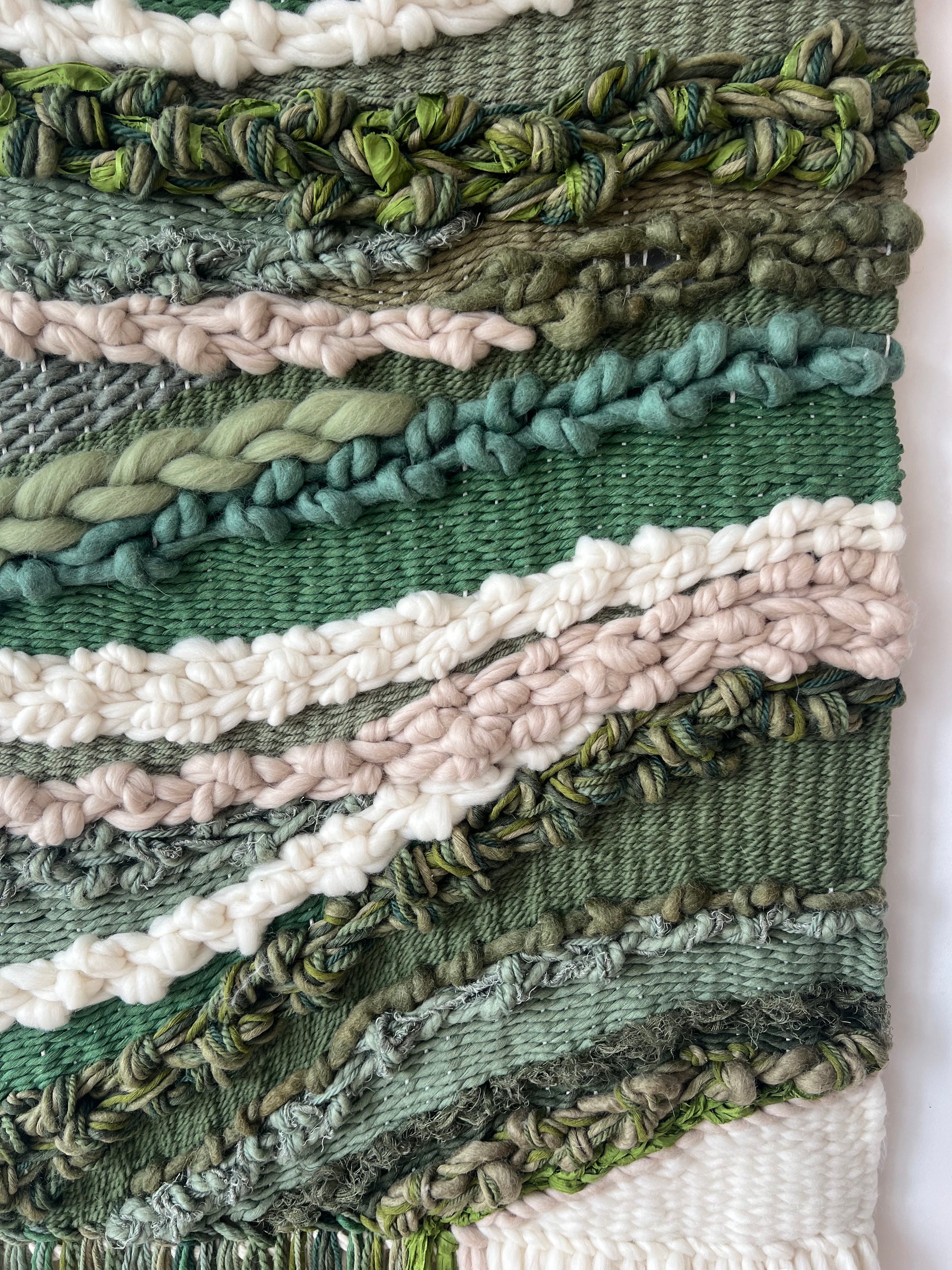 Close-up of soft wool fibers in green, cream and beige tones, showing woven texture of the wall hanging.