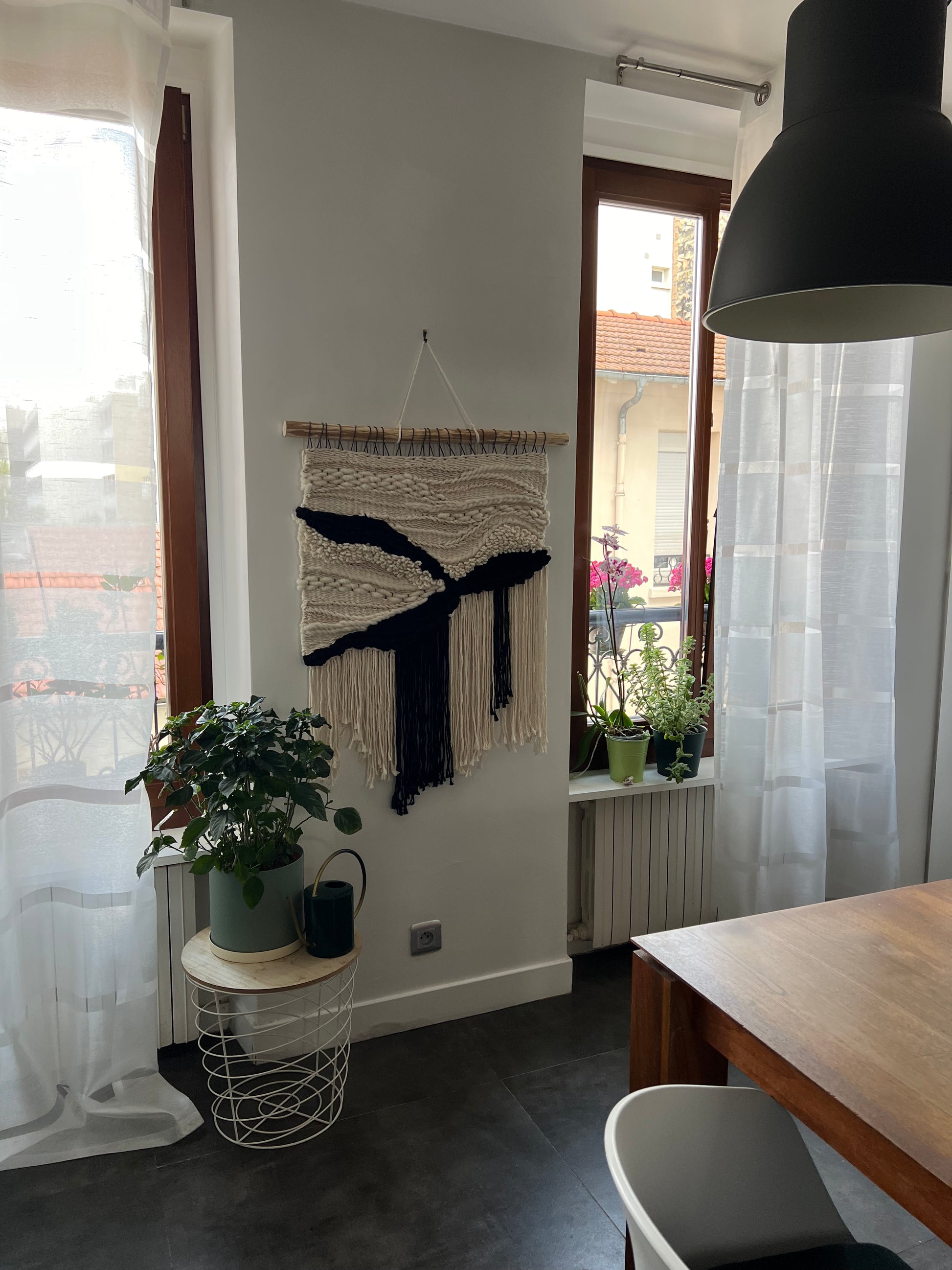 Textured woven wall panel in cream and dark yarn hanging between two kitchen windows with plants