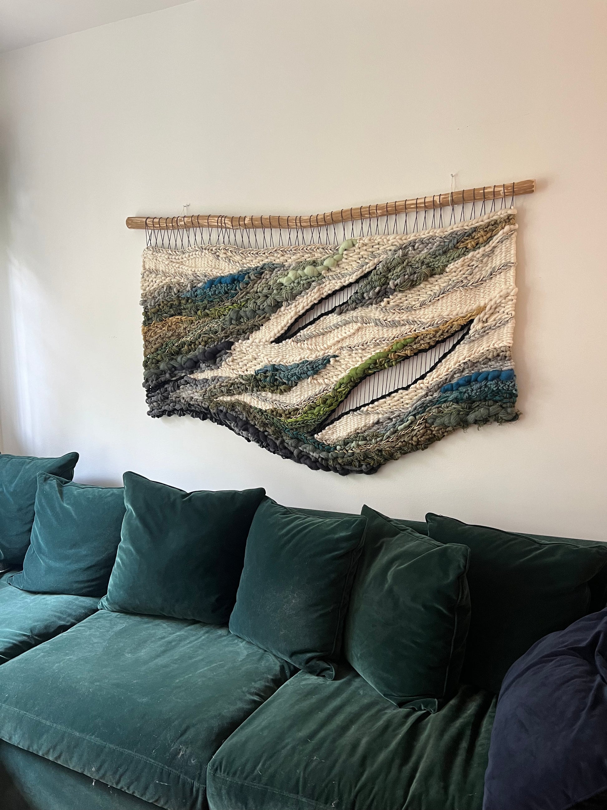 Side view of handmade woven wall art with textured layers of wool and fiber in natural greens, blues, and neutrals