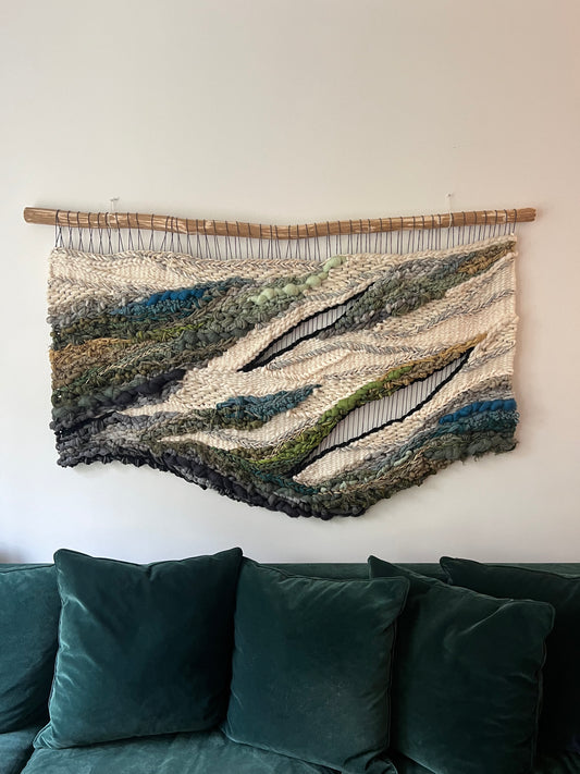 Large handwoven wall hanging in earthy tones of green, blue, and cream, styled above a living room sofa as boho textile art decor