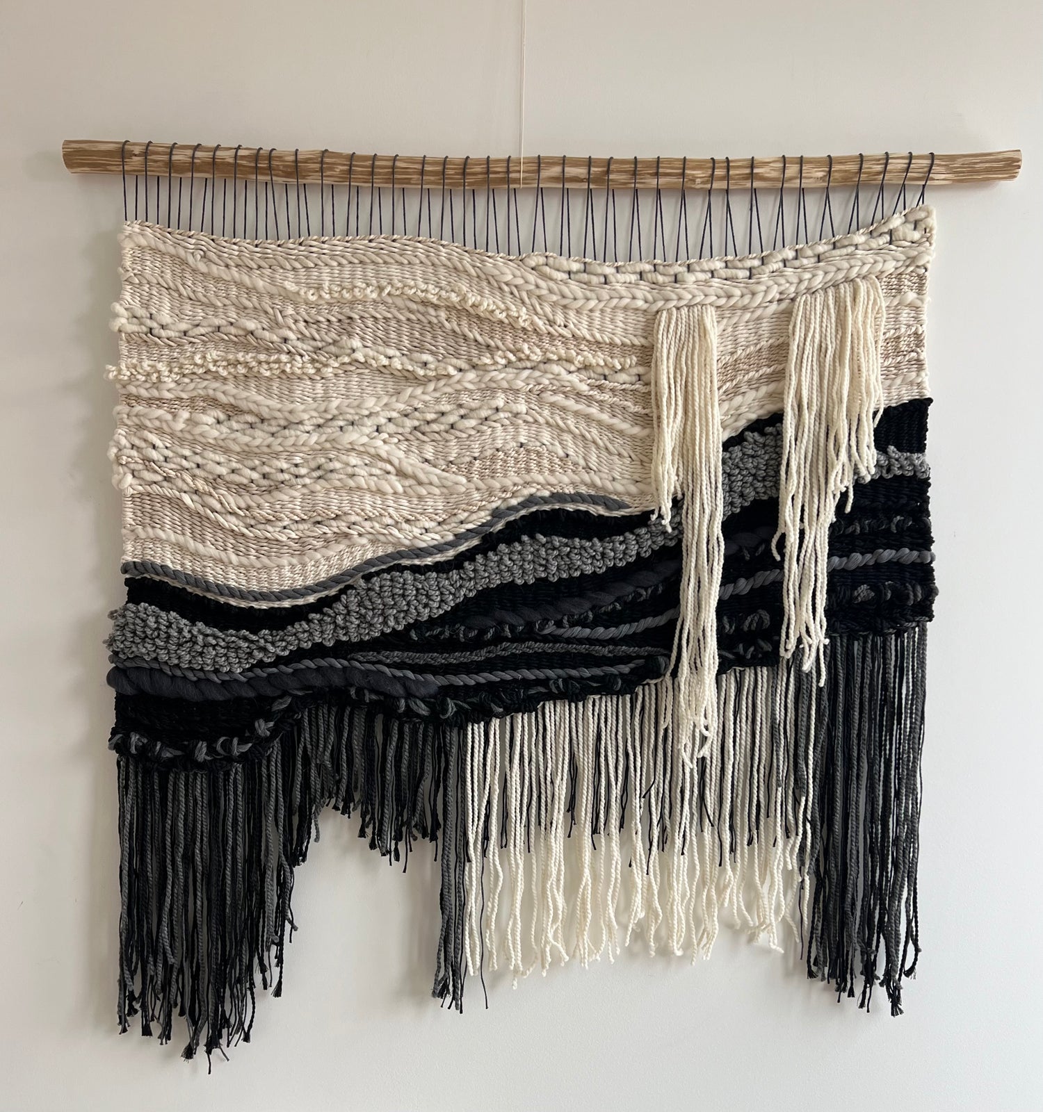 Full view of black and cream woven wall hanging with fringe displayed on plain white wall