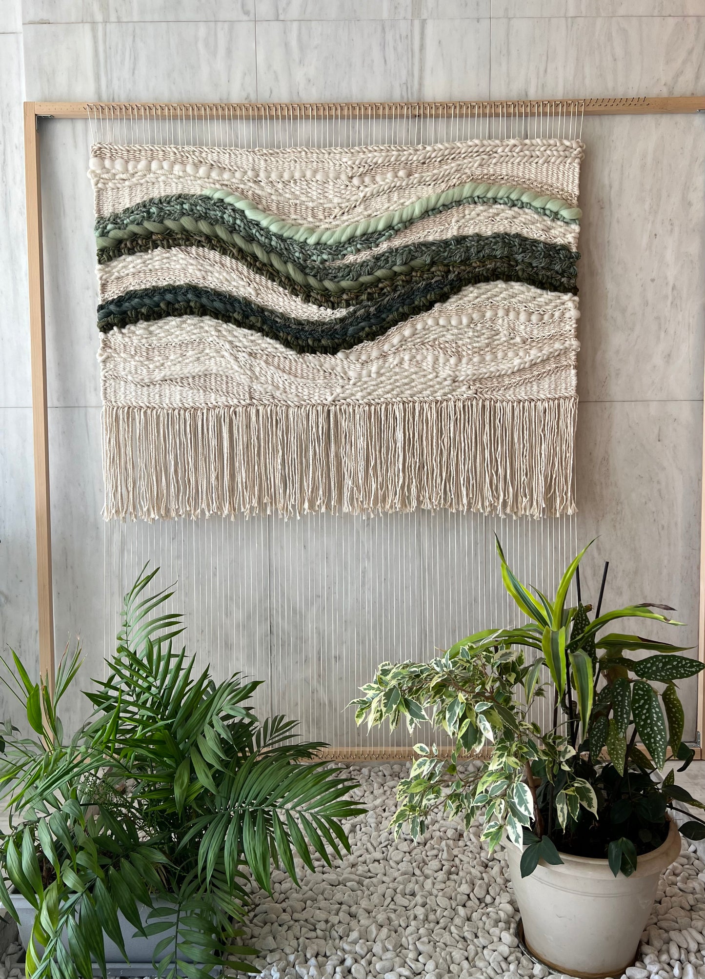 Work in progress of square woven wall hanging on wooden loom, showing cream textured weaving and green waves