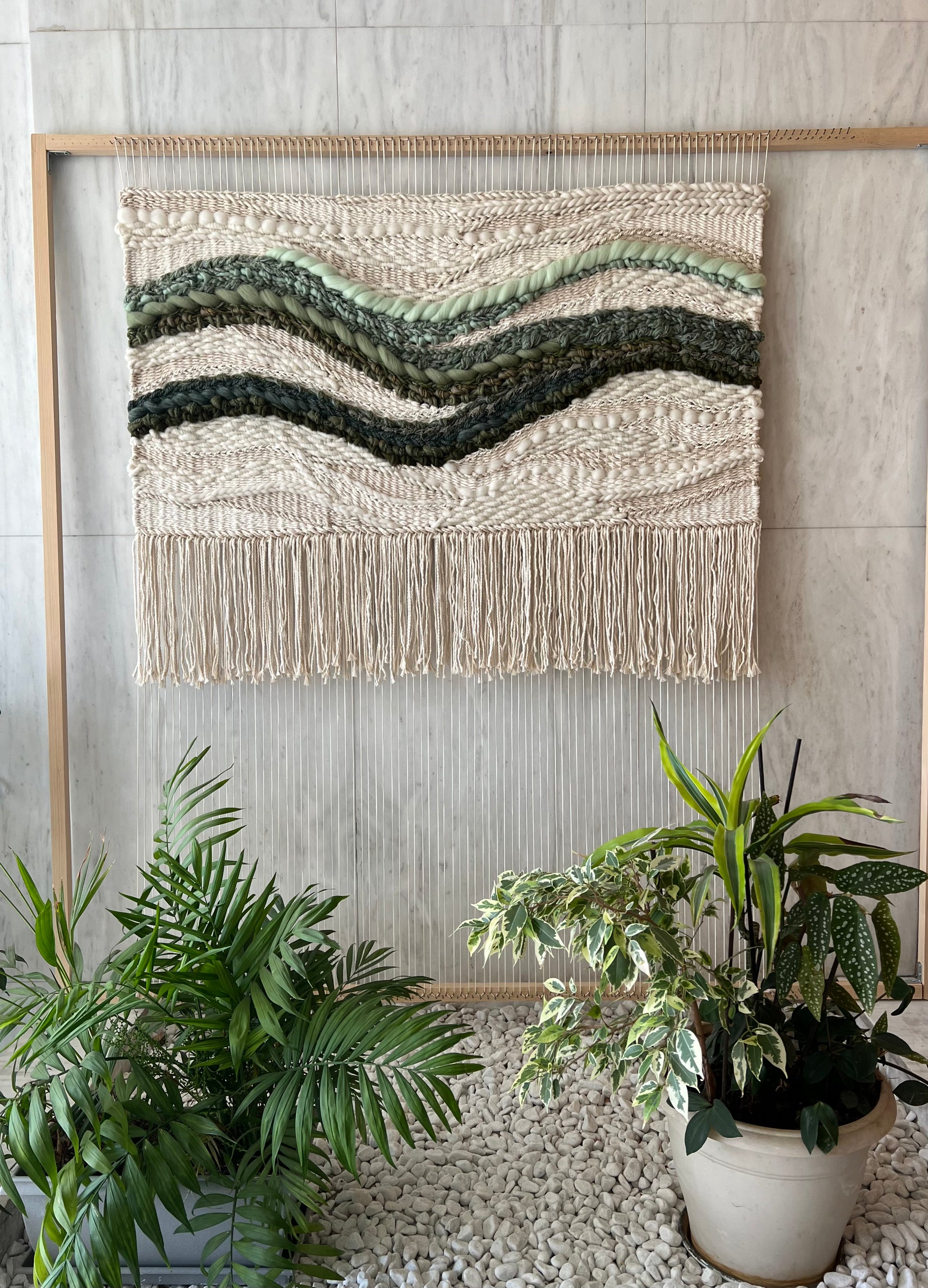 Work in progress of square woven wall hanging on wooden loom, showing cream textured weaving and green waves