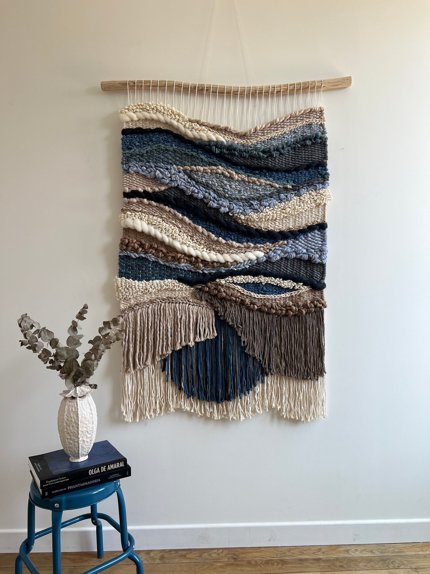 Large handwoven wall hanging in blue, cream, and earthy brown tones, styled in a modern interior with vase and books — boho textile art decor